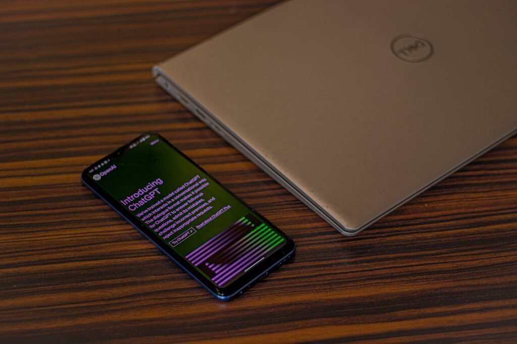 A smartphone displaying ChatGPT app beside a closed laptop on a wooden table.